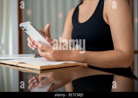 Cropped image of Thai woman using smartphone sitting at table with book in cafe Stock Photo