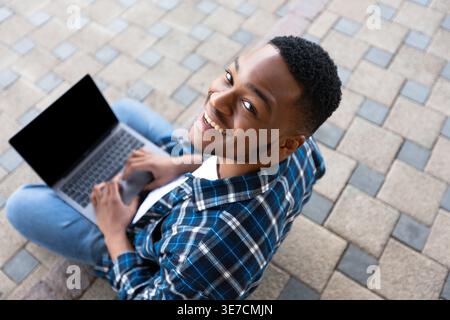 Black man with laptop looking up sitting on sidewalk in park Stock Photo