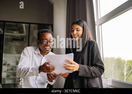 Black businessman in eyeglasses reviewing contract with mixed-race female colleague while standing together in office Stock Photo