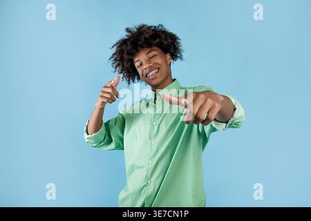 Smiling Black male with curly hair and dental braces wearing long-sleeved shirt pointing fingers forward standing with head tilted sideways  in fro... Stock Photo