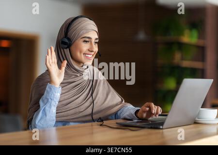 Smiling arab woman having video call while resting at cafe, using laptop and headset, empty space. Cheerful young lady in hijab waving at laptop sc... Stock Photo