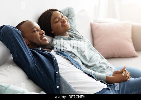 Black husband and wife holding hands while sitting with closed eyes on sofa in living room at home Stock Photo
