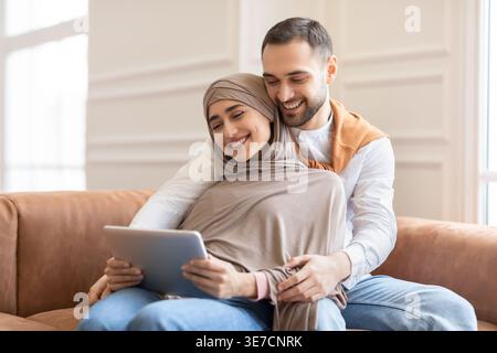 Arab Muslim woman in hijab and caucasian husband using tablet and hugging while sitting on sofa at home Stock Photo