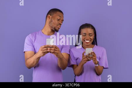 Black couple using smartphones while standing against purple background in studio Stock Photo