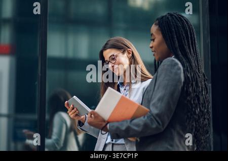 Black businesswoman with braids holding book talking to caucasian female colleague showing tablet standing outside office building during daytime Stock Photo