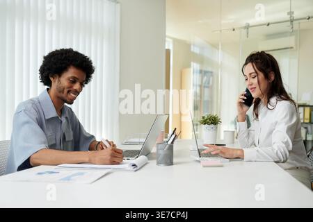 Black businessman with afro taking notes with caucasian female colleague having phone call while sitting together at table with laptop ad financial... Stock Photo