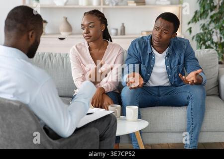 Black man complaining about his wife speaking about their relationship problems while sitting on couch and having marriage counseling with Black ma... Stock Photo