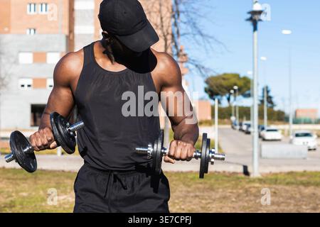 Muscular mixed-race man wearing black tank top and cap lifting dumbbells while standing in park on sunny day Stock Photo