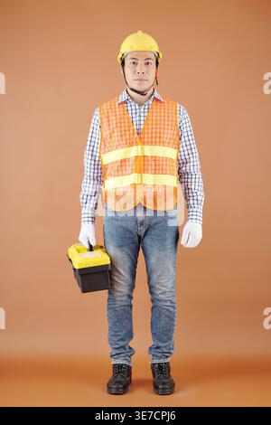 Asian male handyman wearing vest, hardhat and gloves carrying toolbox standing against brown background in studio Stock Photo