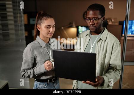 Black male programmer wearing eyeglasses working on laptop standing beside caucasian female colleague in office at night Stock Photo