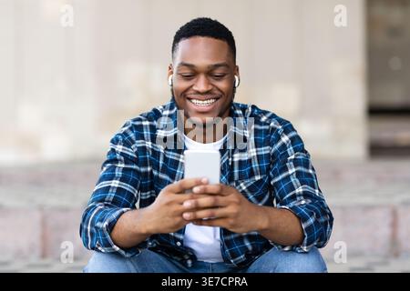 Black man using smartphone listening to music with earphones sitting against concrete wall outdoors Stock Photo