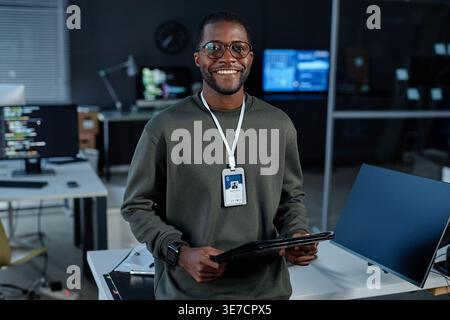 Smiling Black male programmer wearing eyeglasses holding folder while standing against table with computers in office at night Stock Photo
