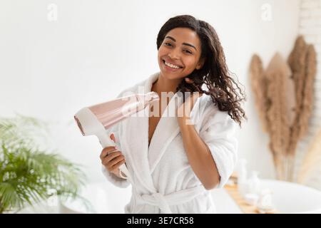 Black woman in bathrobe blow drying hair standing in bathroom at home Stock Photo