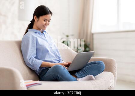 Smiling Asian woman using laptop sitting on couch with crossed legs in living room at home Stock Photo