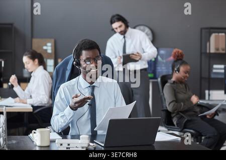 Black male broker wearing eyeglasses and headset talking to customer while sitting at his desk with laptop in office Stock Photo