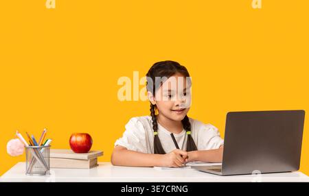 Asian schoolgirl with braids taking notes and looking at laptop sitting at table with books against yellow background in studio Stock Photo