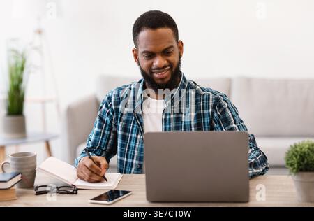 Black businessman taking notes while looking at laptop sitting at table in living room at home Stock Photo
