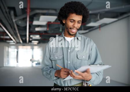 Black male contractor in gray uniform taking notes while standing and inspecting construction site Stock Photo