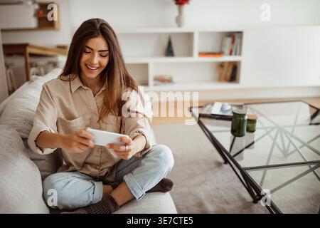 Cheerful Hispanic woman having fun playing games on her smartphone while sitting with crossed legs on sofa in living room at home Stock Photo