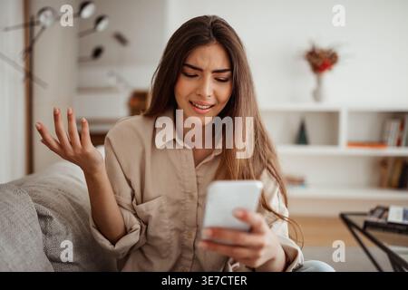 Confused Hispanic woman using smartphone sitting on sofa in living room at home Stock Photo