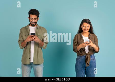 Smiling Asian couple using smartphones while standing against blue background in studio Stock Photo