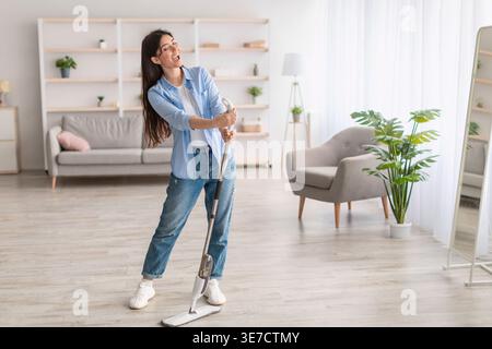 Middle Eastern woman with closed eyes holding mop singing while standing in living room at home Stock Photo
