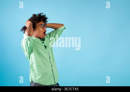 Shocked Black male with curly hair and dental braces looking away holding head with both hands standing with mouth opened in front of blue backgrou... Stock Photo