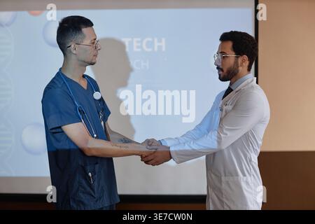 Mixed-race and Middle Eastern male doctors wearing eyeglasses shaking hands during medical presentation while standing together near projector scre... Stock Photo