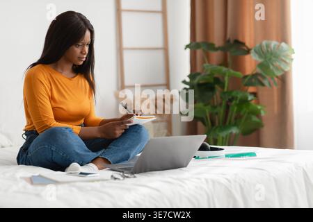 Back female student writing in notebook studying and looking at laptop sitting with crossed legs on bed in bedroom at home Stock Photo