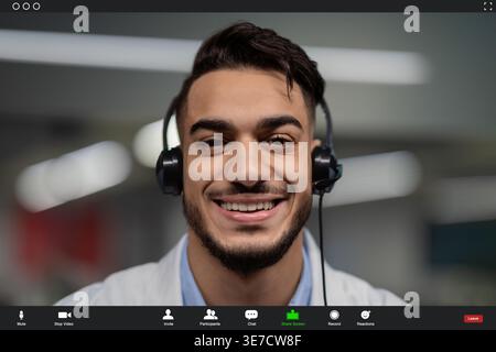 Smiling Arab male doctor wearing lab coat and headset having video call with patient in office Stock Photo