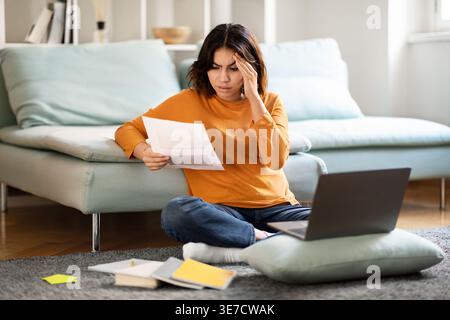 Stressed Hispanic woman touching her forehead reading papers sitting on floor with laptop and books in front of bed in bedroom at home Stock Photo