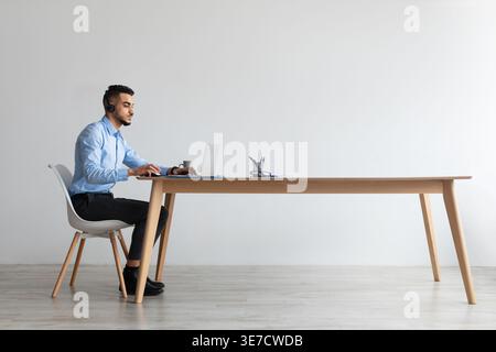 Serious Middle Eastern man wearing headphones working on laptop sitting at tablewith coffee cup in office Stock Photo