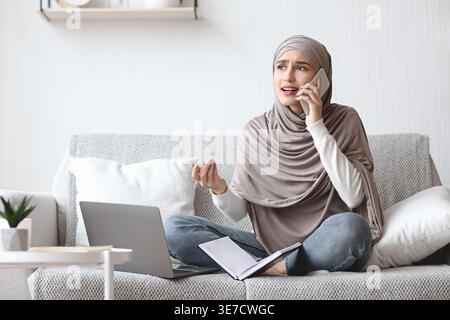 Disappointed Arab Muslim woman in hijab gesturing hand having phone call sitting with legs crossed on couch with laptop and notebook at home Stock Photo
