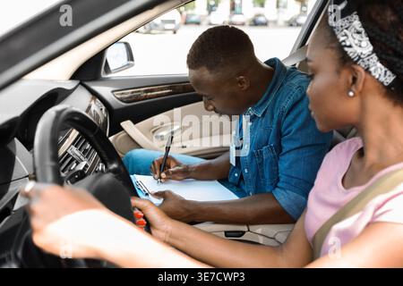 Black male driving instructor writing notes on evaluation sheet of female student while sitting inside car after driving lesson Stock Photo