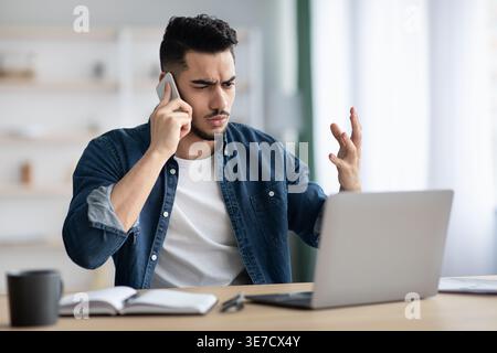 Middle Eastern businessman having phone call and looking at laptop sitting at table with notebook at home Stock Photo