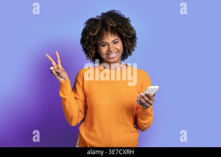 Black woman with afro showing peace hand sign holding smartphone standing against purple background in studio Stock Photo