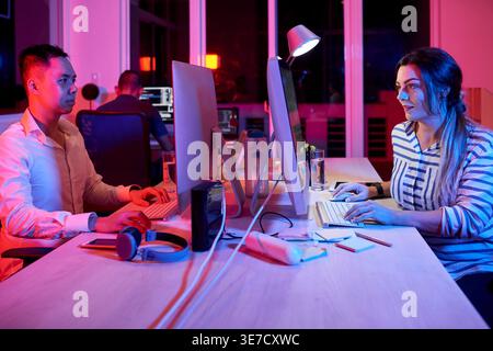 Team of software developers working on project sitting at desks with computers in office at night Stock Photo
