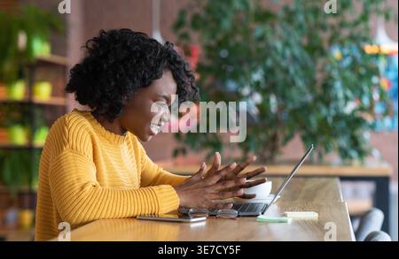 Annoyed Black woman with open hands looking at laptop sitting at table in cafe Stock Photo