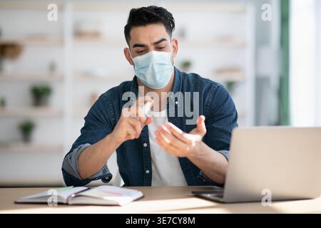 Middle Eastern man wearing protective face mask using disinfectant for hands sitting at table with laptop at home Stock Photo