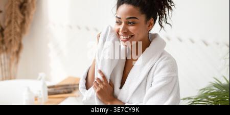 Black woman in bathrobe drying face with towel looking away while sitting in bathroom at home Stock Photo