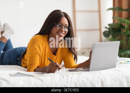 Black female student wearing eyeglasses taking notes studying and using laptop lying on bed in bedroom at home Stock Photo