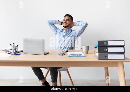 Relaxed Middle Eastern man with closed eyes and hands behind head sitting at table with laptop in office Stock Photo