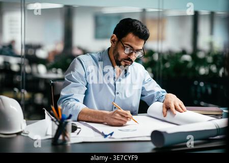 Middle Eastern male architect with eyeglasses sketching on blueprint plan sitting on desk in office Stock Photo