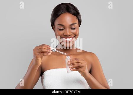Black woman wrapped in towel holding bottle and dropper of skin serum standing against gray background in studio Stock Photo