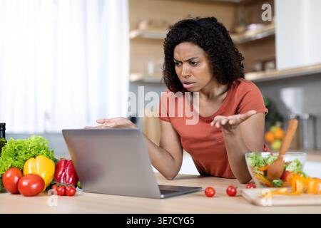 Black woman having video call on laptop gesturing hands standing and leaning on kitchen counter with vegetables at home Stock Photo