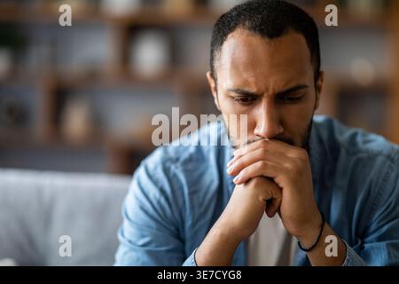Stressed Black man leaning mouth on clasped hands sitting on sofa in living room at home Stock Photo