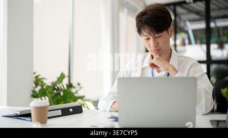 Focused Asian male colleague leaning chin on hand working on laptop sitting at desk in office Stock Photo