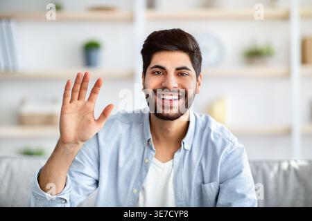 Smiling Arab man with beard waving hand while sitting on sofa in living room at home Stock Photo