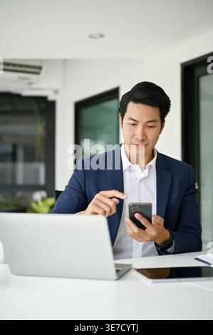 Asian businessman using smLifestylephone sitting at table with laptop in office during daytime Stock Photo