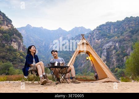Cheerful Chinese couple looking away sitting on folding chairs having breakfast near mountains during camping Stock Photo